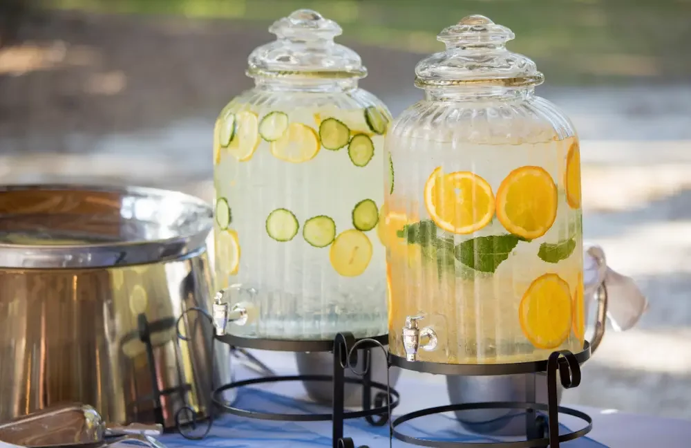 Two glass beverage dispensers filled with water infused with cucumbers and citrus slices, on a tray outdoors.