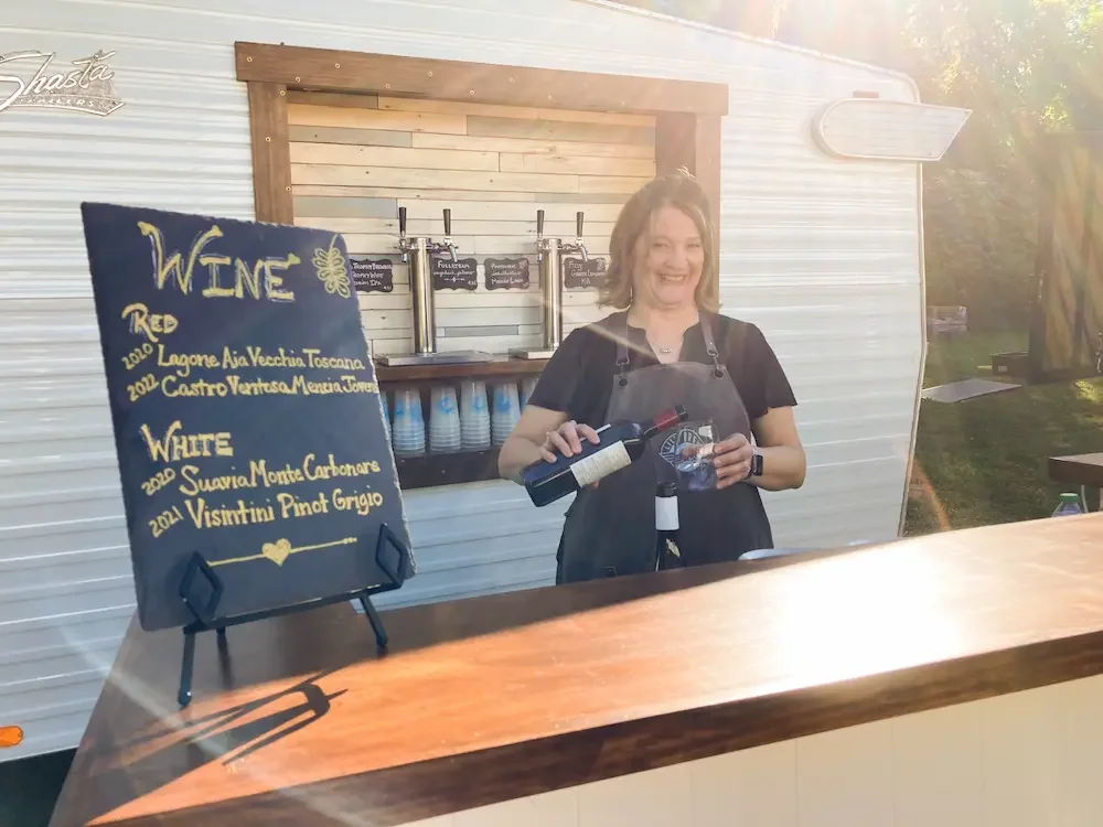 A woman standing behind a wooden bar counter, pouring red wine into a glass, with a blackboard sign displaying different wines, and a mobile bar setup behind her.