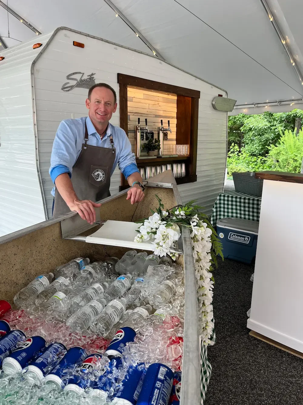 A mobile bartender with apron at a beverage stand with bottled water, soda cans, and flowers in a tented outdoor setting.