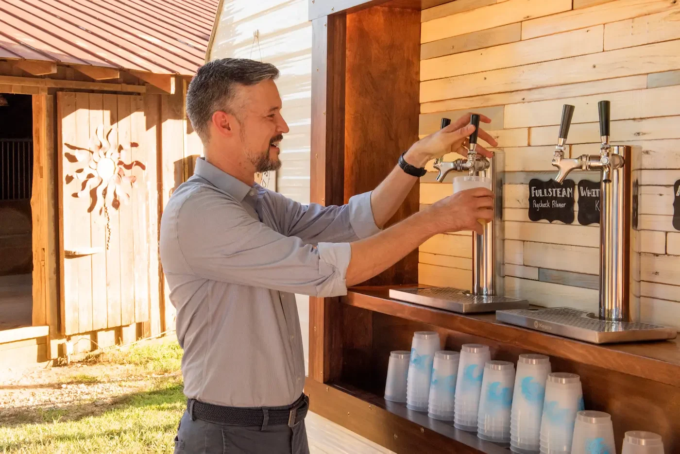 A man in a gray shirt pouring beer from a tap at an outdoor bar with wooden walls.