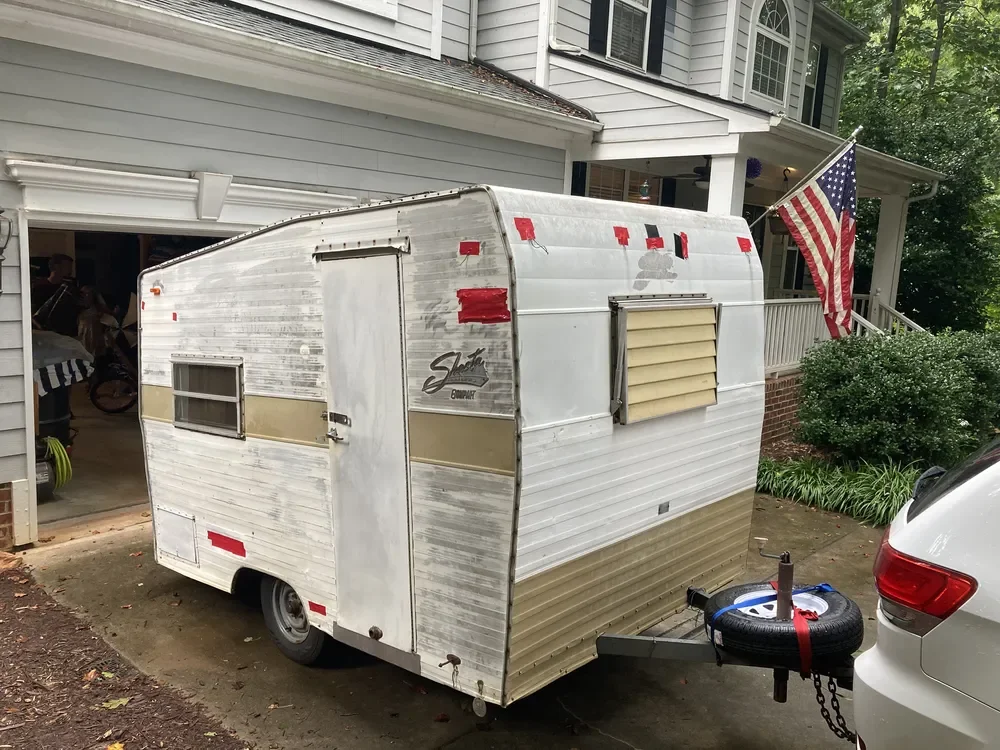 A small travel trailer parked in a residential driveway near a white house with an American flag. The trailer is white and tan with a window and a louvered vent, showing signs of wear and some red tape.