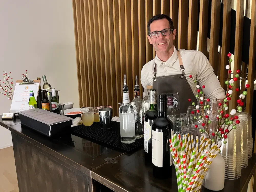 A smiling bartender standing behind a bar with various bottles of alcohol, colorful straws, and holiday decorations including red and green berries.