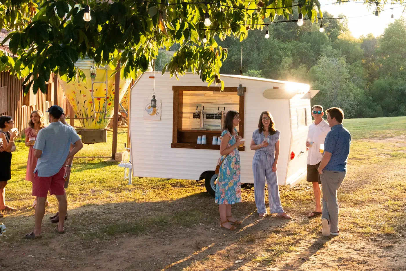 Group of people socializing outdoors near the vintage tap trailer with a wooden window frame, decorated with string lights and a large decorative vase, during sunset.