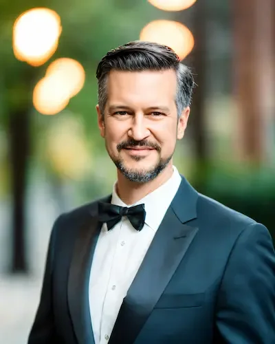 A man in a tuxedo with a bow tie smiling outdoors during the evening with blurred lights in the background.