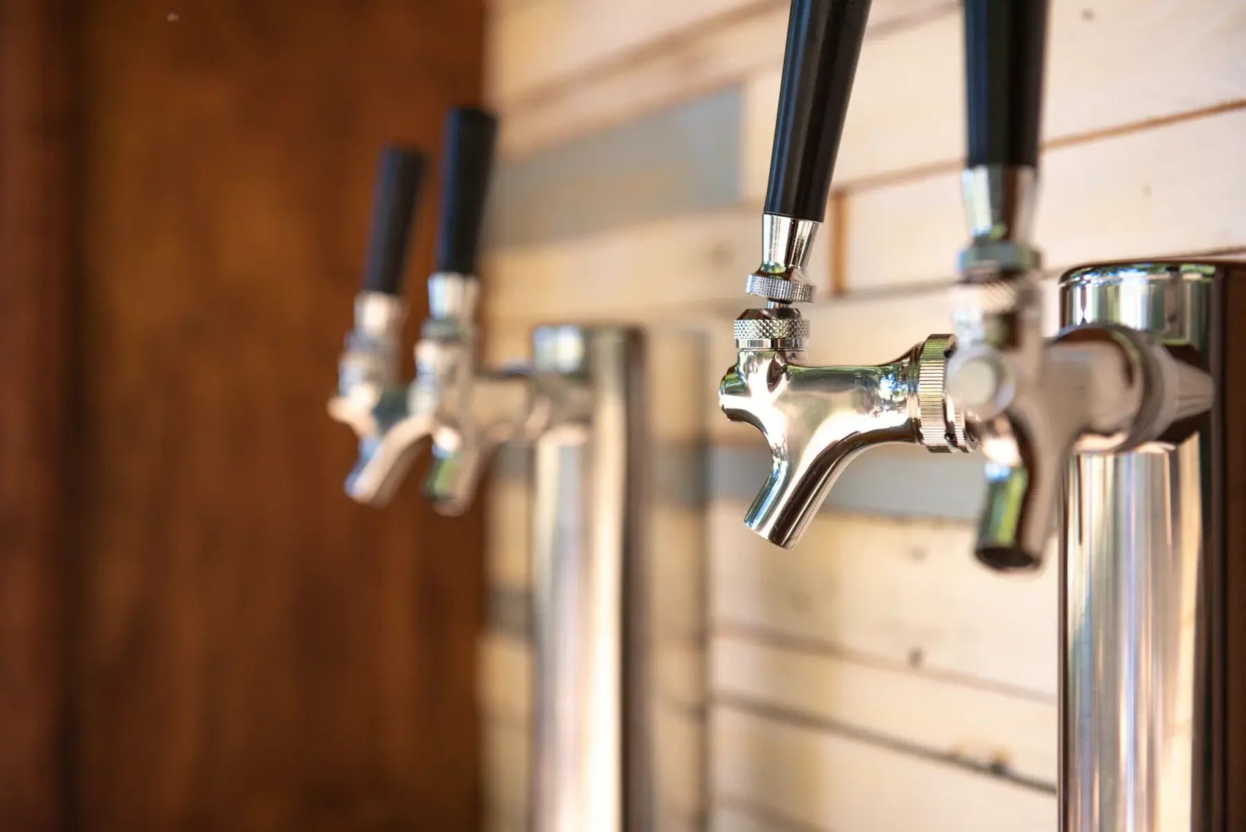 Close-up of chrome beverage taps with black handles mounted on a wooden wall.