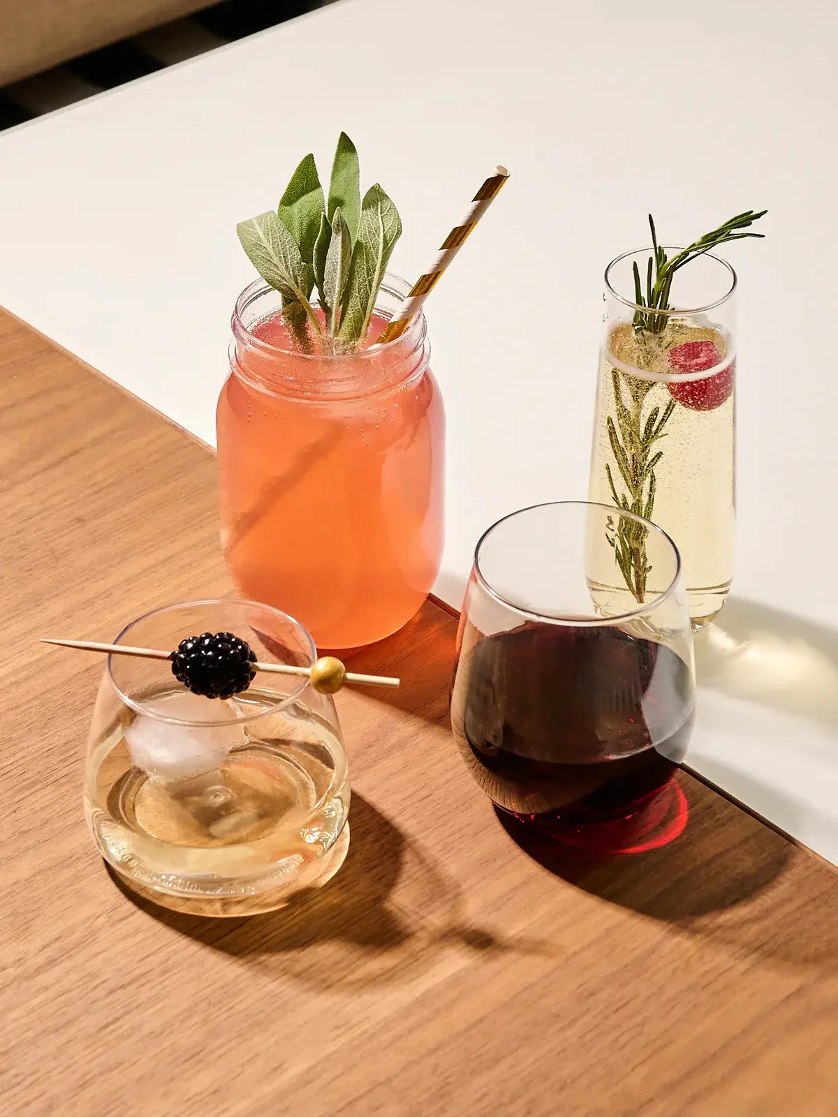 Four drinks on a wooden surface, including a pink drink garnished with sage leaves in a mason jar, a clear drink with a blackberry and skewer, a dark red wine, and a bubbly beverage with herbs and a cherry inside a tall glass.