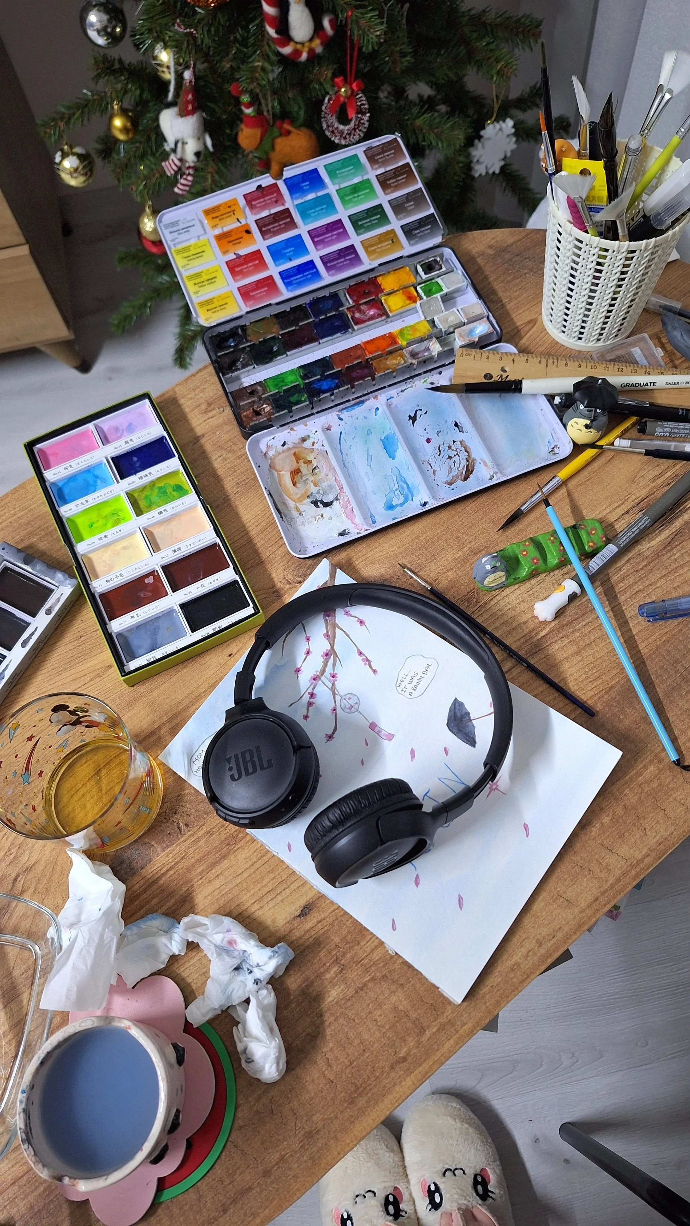 cluttered desk with two different paint pallets, papers, headphones strewn about on a desk