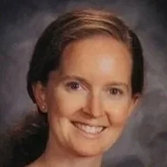 Smiling woman with brown hair in a ponytail, wearing an orange and white striped top, against a dark background.