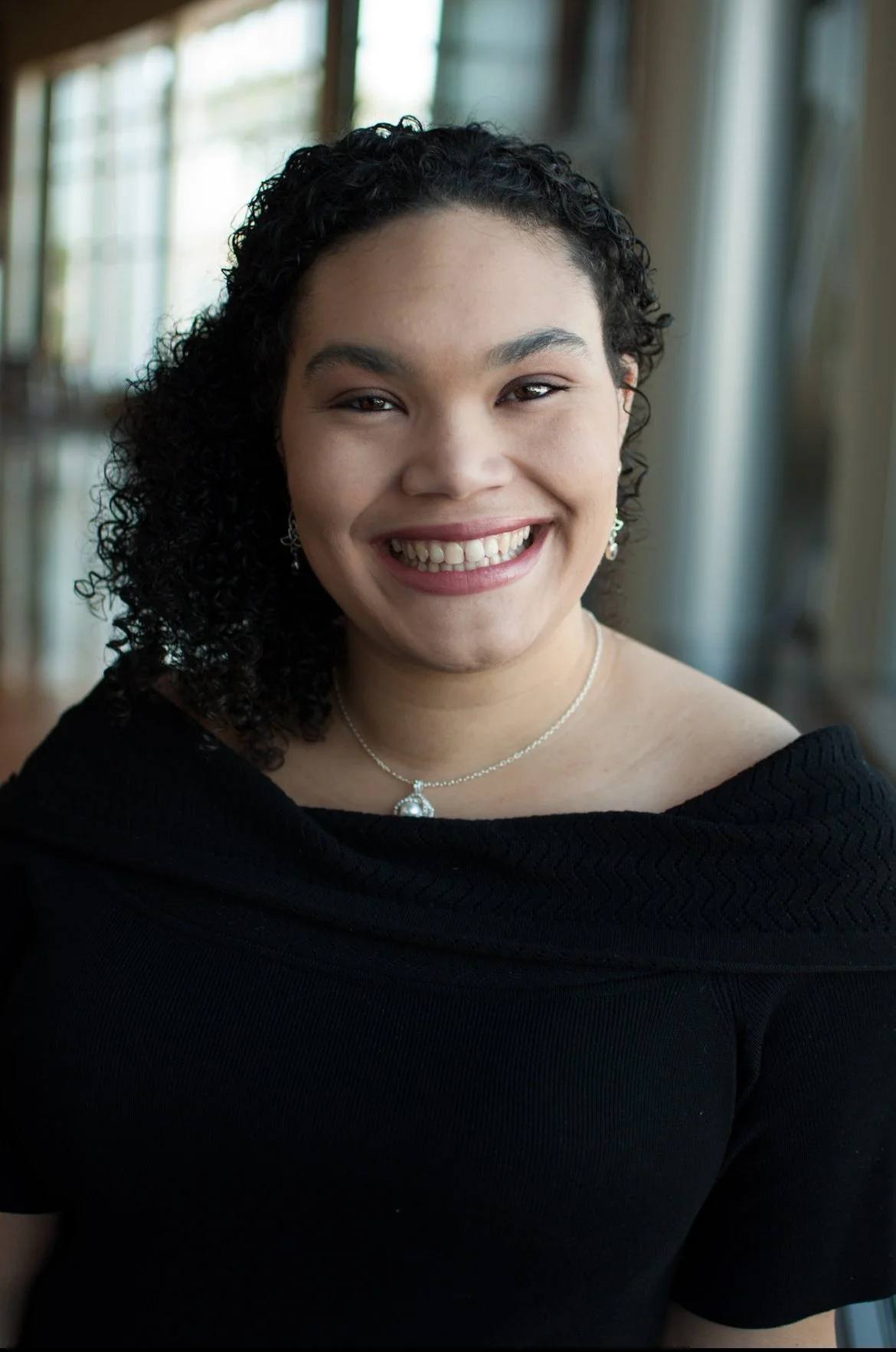 A young woman with curly black hair and a bright smile, wearing a black top, silver earrings, and a silver necklace with a pendant, in a room with large windows and natural light.