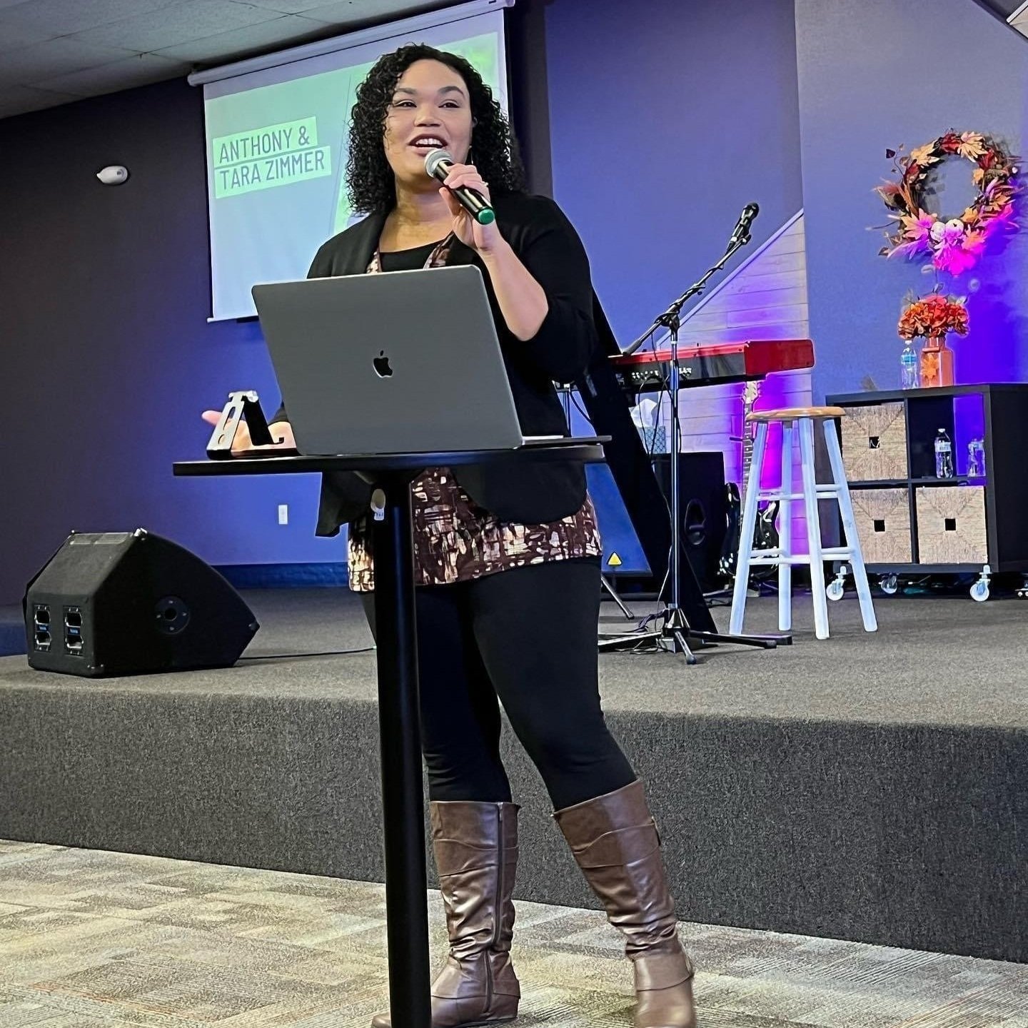 Woman speaking at an event with a microphone, standing at a podium with a laptop, stage setup with musical instruments and decorations.