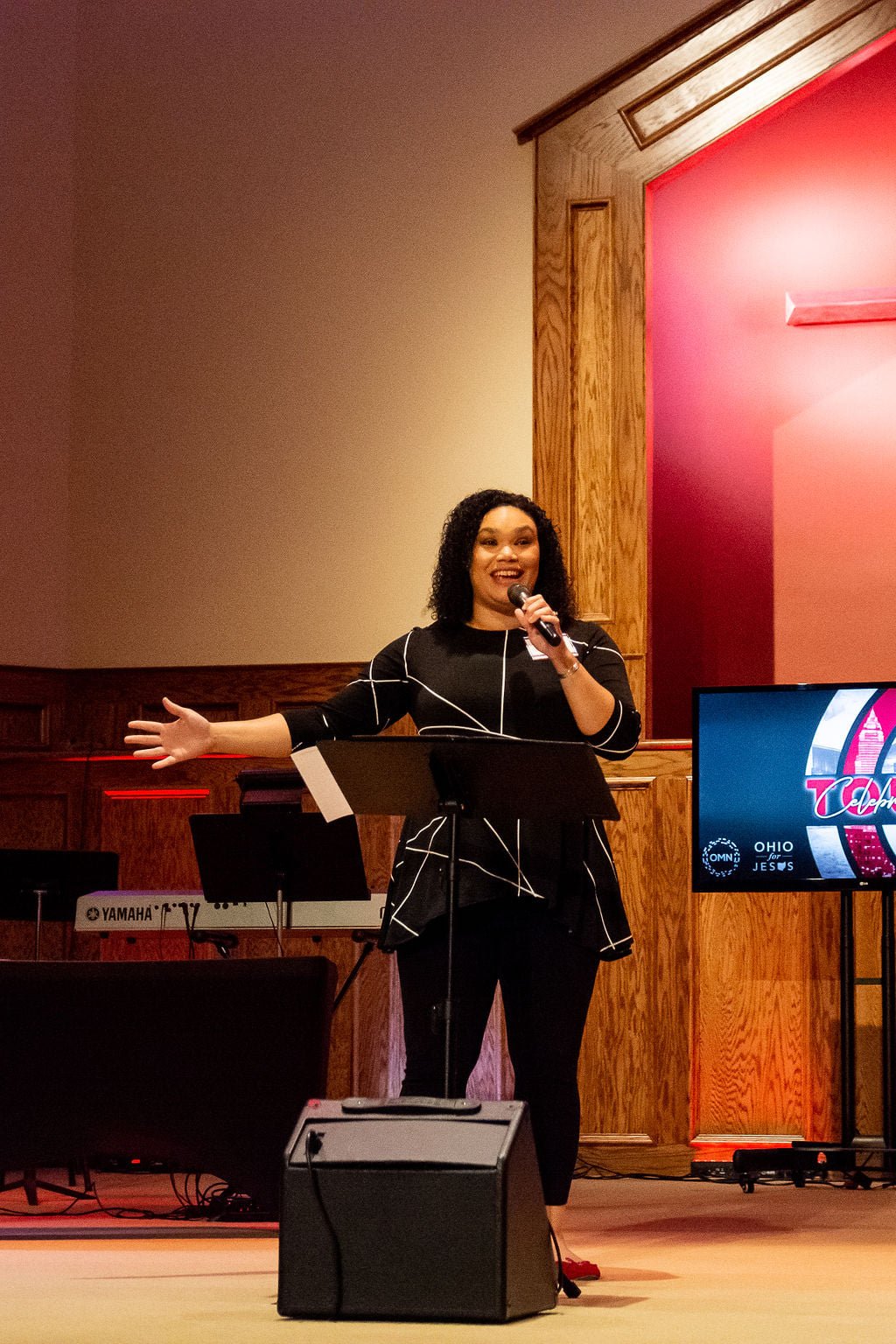 A woman with curly hair singing or speaking into a microphone on stage, with a keyboard, monitor, and a large screen displaying a promotional design in the background.