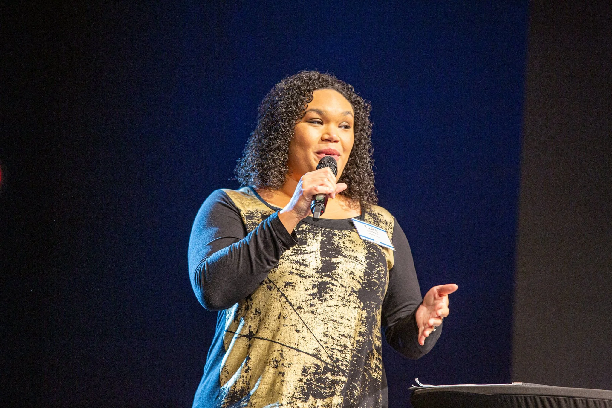 A woman with curly hair, wearing a black and gold patterned dress, speaking into a microphone on stage with a dark background.