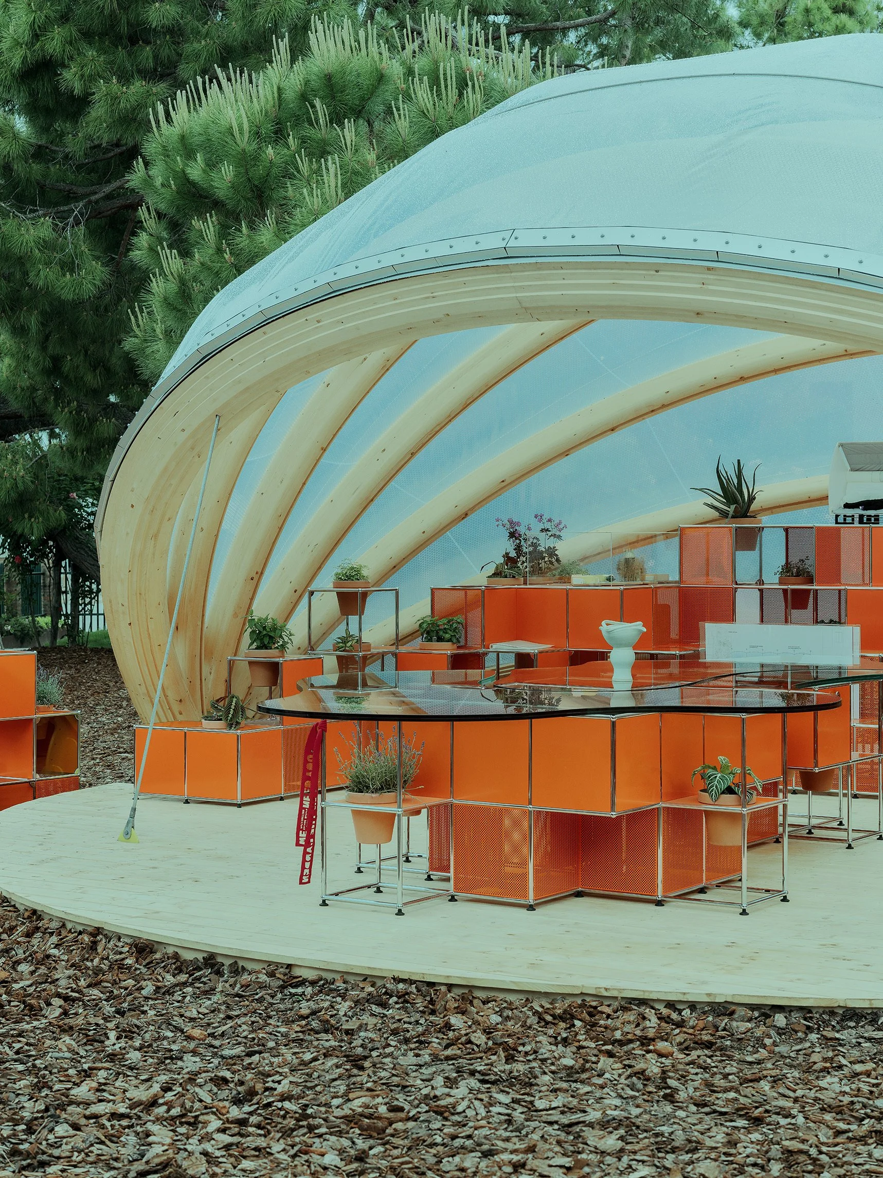 Outdoor Exhibit at Giardini della Marinaressa with a curved roof, wooden arches, and orange USM Modular furniture with potted plants and greenery inside. Outside, there are trees and a wood chip ground covering.
