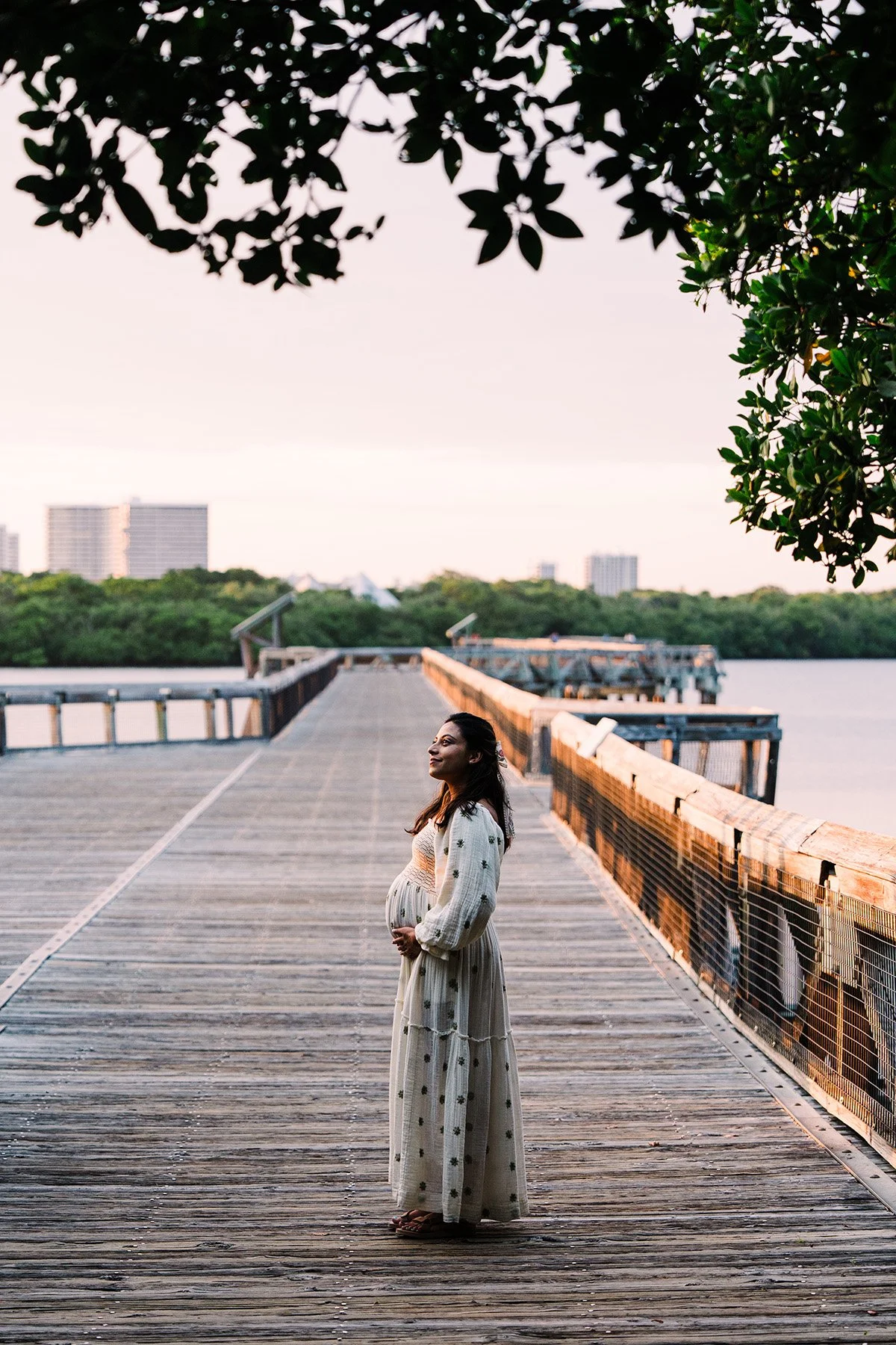 A pregnant woman in a long, white, floral dress standing on a wooden pier by the water, smiling and looking to the side during sunset with trees and city buildings in the background.
