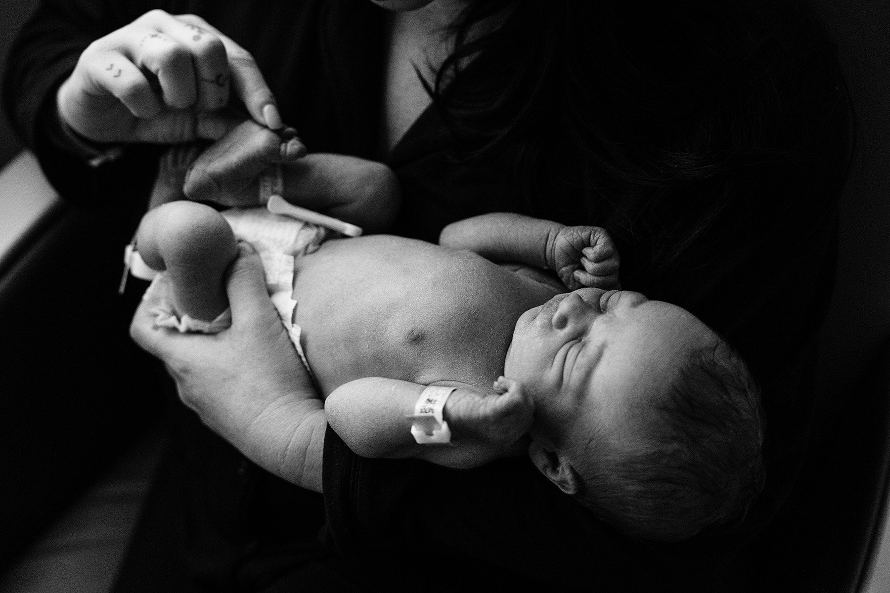 Black and white photo of a newborn baby lying in someone's arms, with a hospital wristband on the baby's wrist, and a hand holding a small object or syringe near the baby's head.