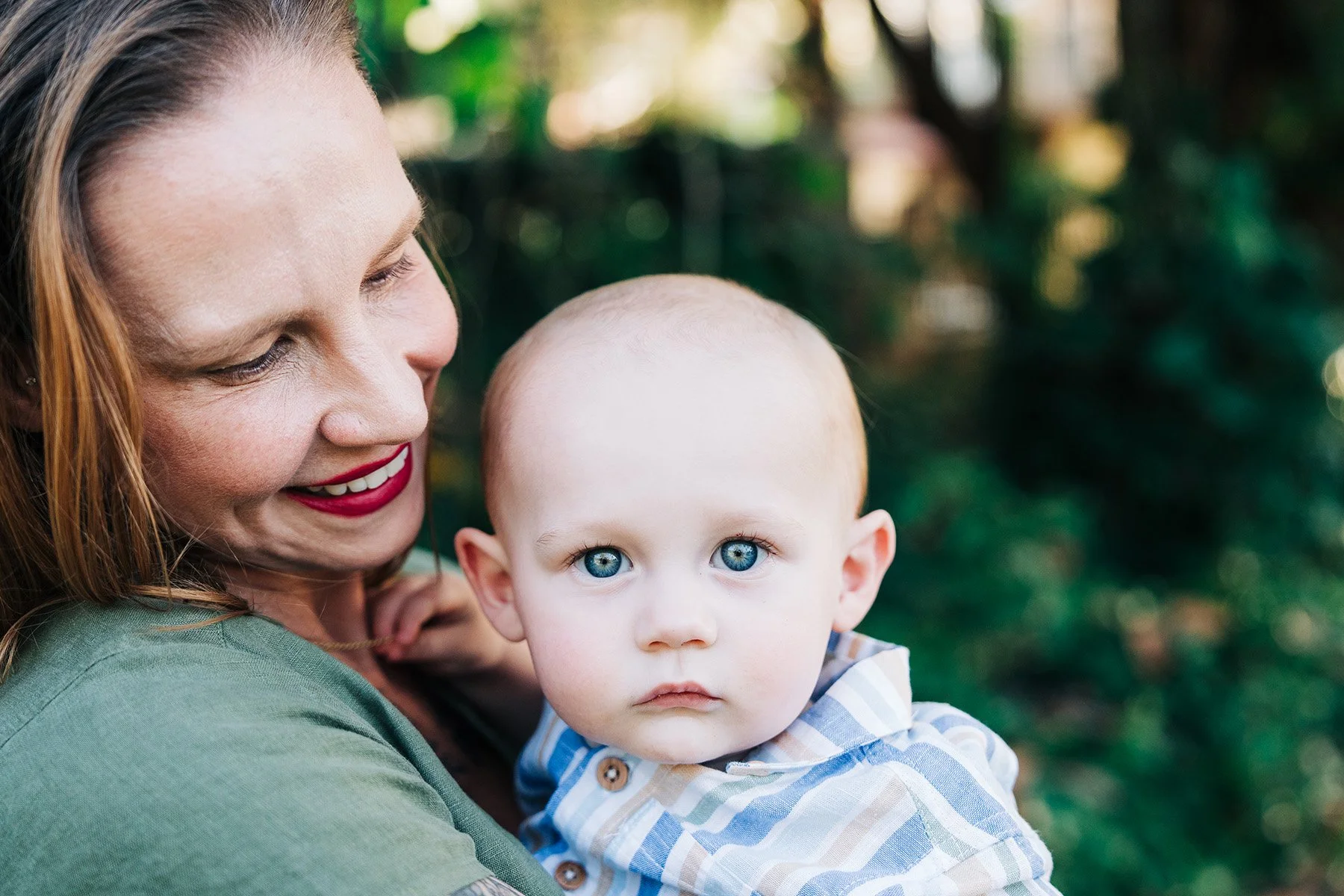 A woman with red hair, red lipstick, and a green shirt holding a young child with bright blue eyes and light hair outdoors with blurred green foliage in the background.