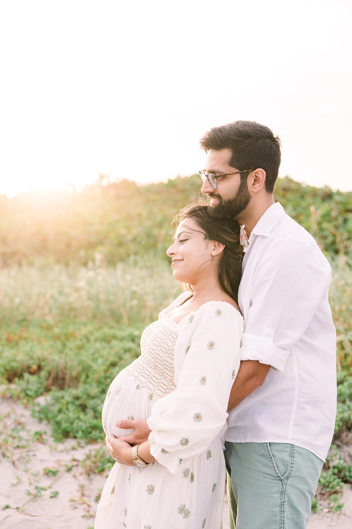 A pregnant woman and a man stand together outdoors during sunset, with the woman leaning back into the man's embrace, both smiling peacefully.