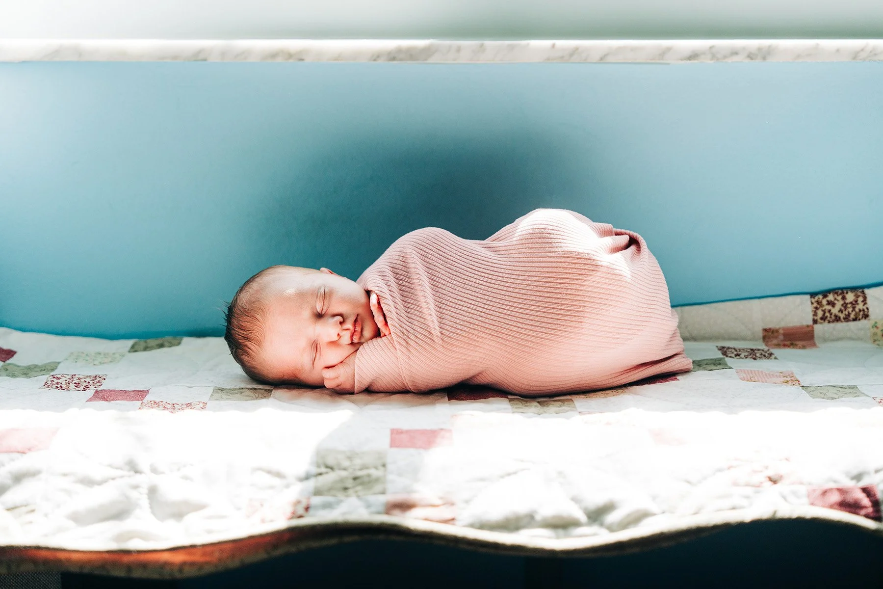 A newborn baby sleeping on a patterned quilt, wrapped in a light pink ribbed onesie, lying on a bed with a quilt and a blue headboard in the background.