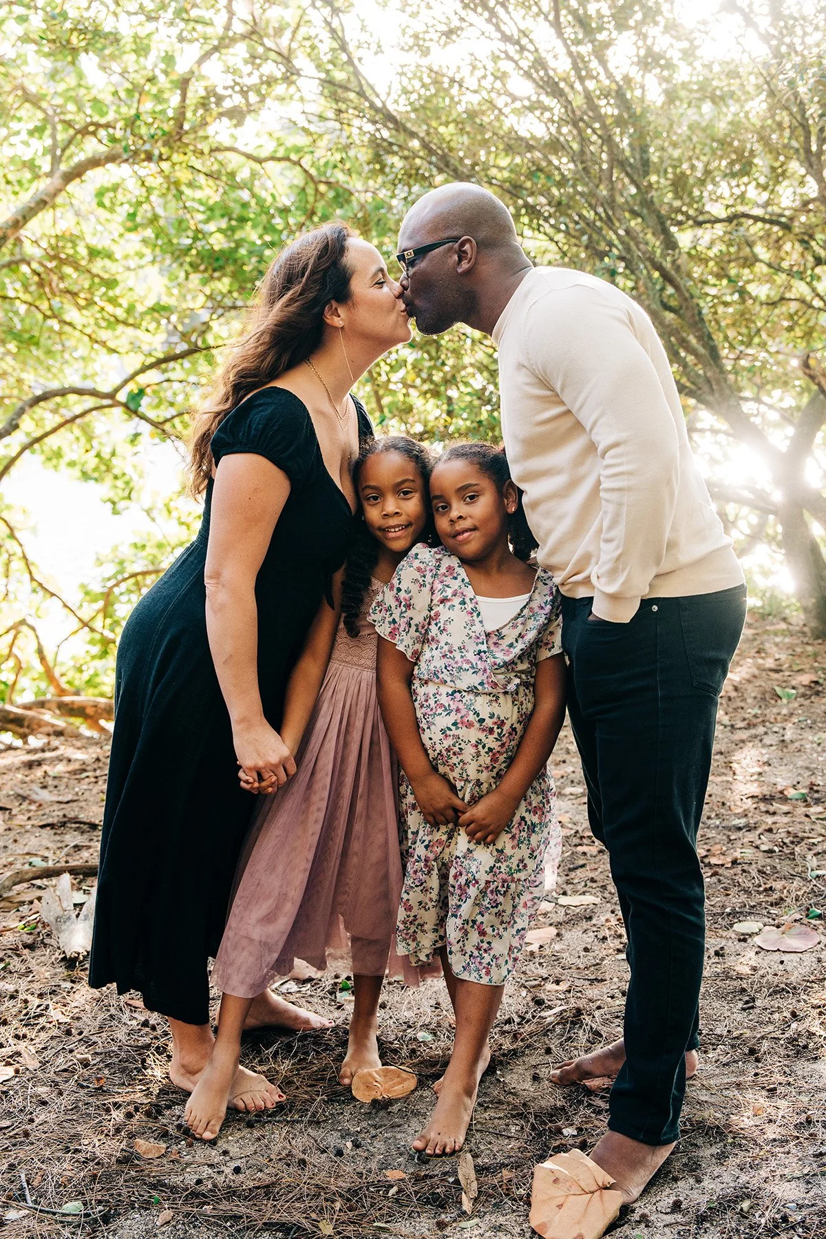 A family of four, including a woman, a man, and two young girls, sharing a kiss outdoors in a wooded area during daytime. They are barefoot and holding hands.