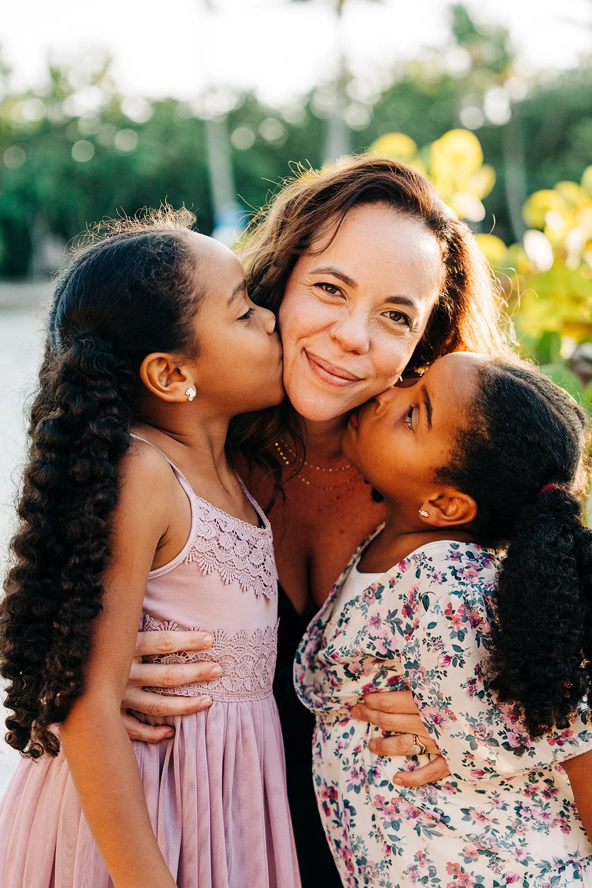 A woman with light brown hair is being kissed on both cheeks by two young girls, one with curly black hair and the other with curly dark brown hair, in an outdoor setting with greenery in the background.