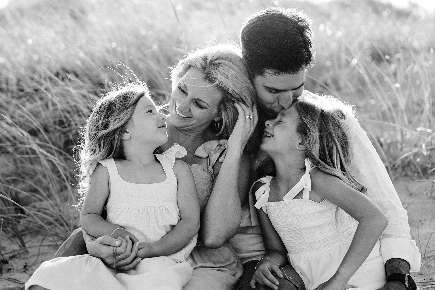 A family of four, including two young girls and their parents, sitting together outdoors in a field, smiling and embracing each other in a joyful moment.
