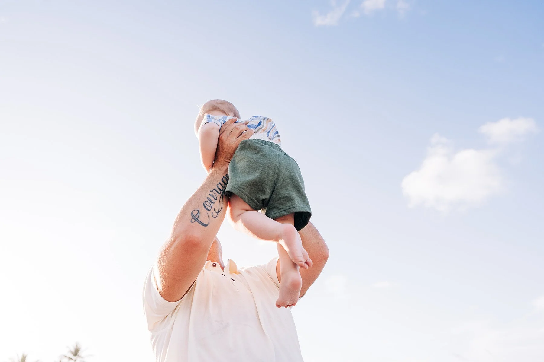 A person holding a young child in the air outdoors against a blue sky with a few clouds, with palm trees visible in the background.
