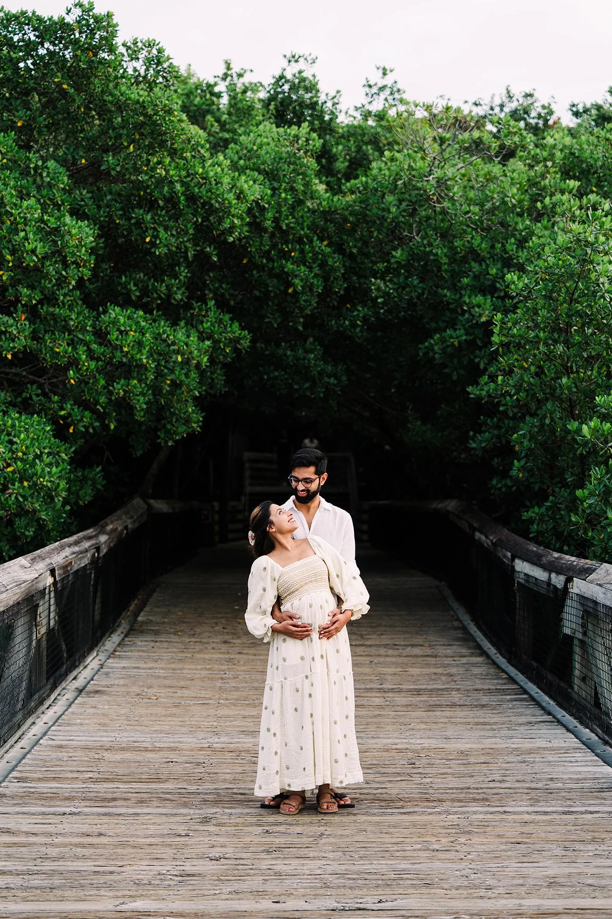 A pregnant woman and a man on a wooden bridge surrounded by green trees, with the woman looking up at the man and holding her belly.