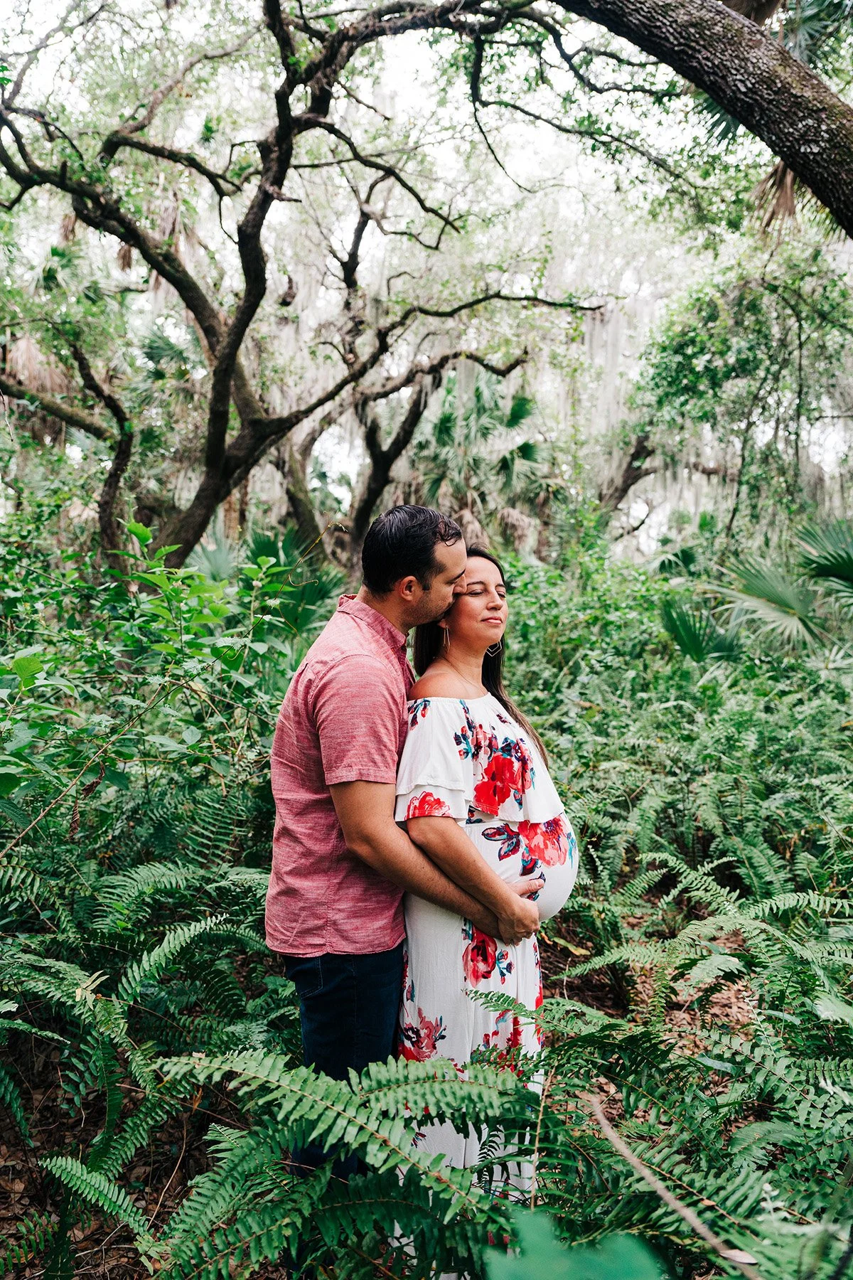A pregnant woman in a white floral dress stands in a lush, green forest with a man in a pink shirt standing behind her, kissing her on the head with his arms around her.