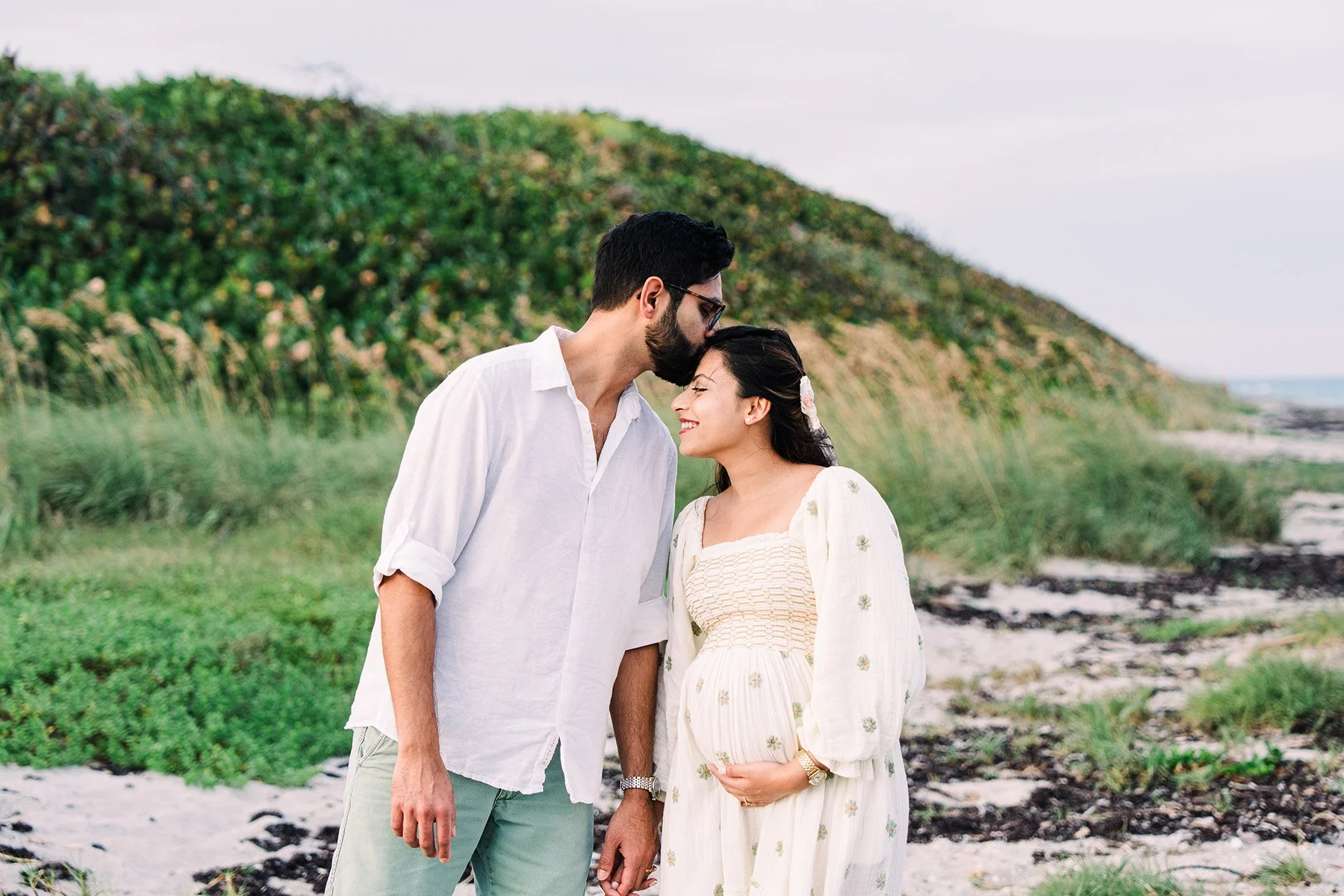 A couple standing closely on a beach with green hills and shoreline in the background. The man gently kisses the woman's forehead as they smile, holding hands.