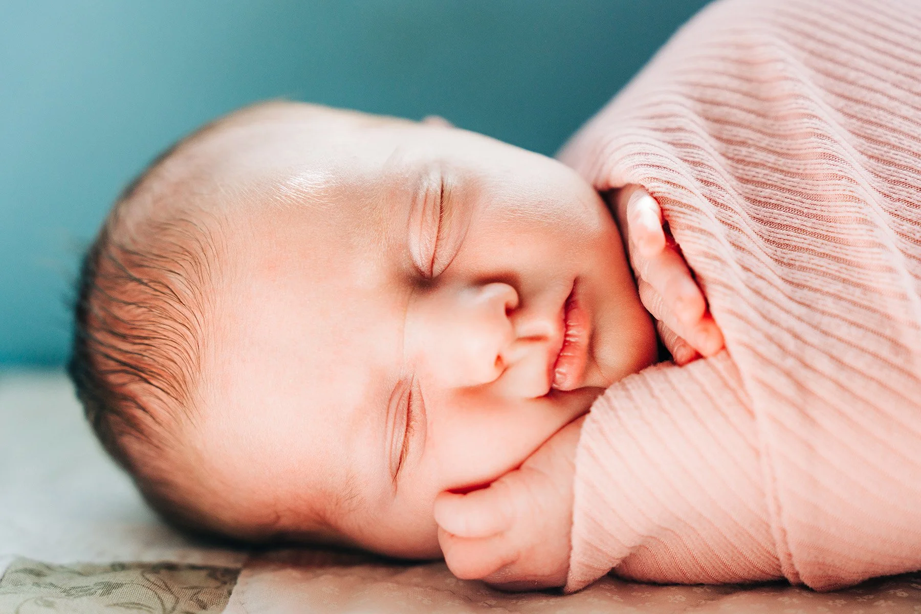Close-up of a sleeping newborn baby with delicate features, resting on a soft surface, wrapped in a light pink, striped fabric.