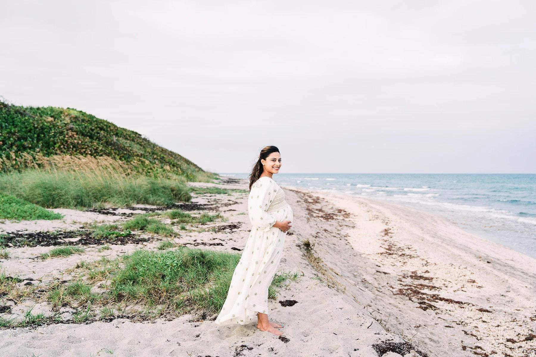 Pregnant woman in a white dress with floral pattern standing barefoot on the beach, smiling and holding her belly, with ocean waves and a cloudy sky in the background.