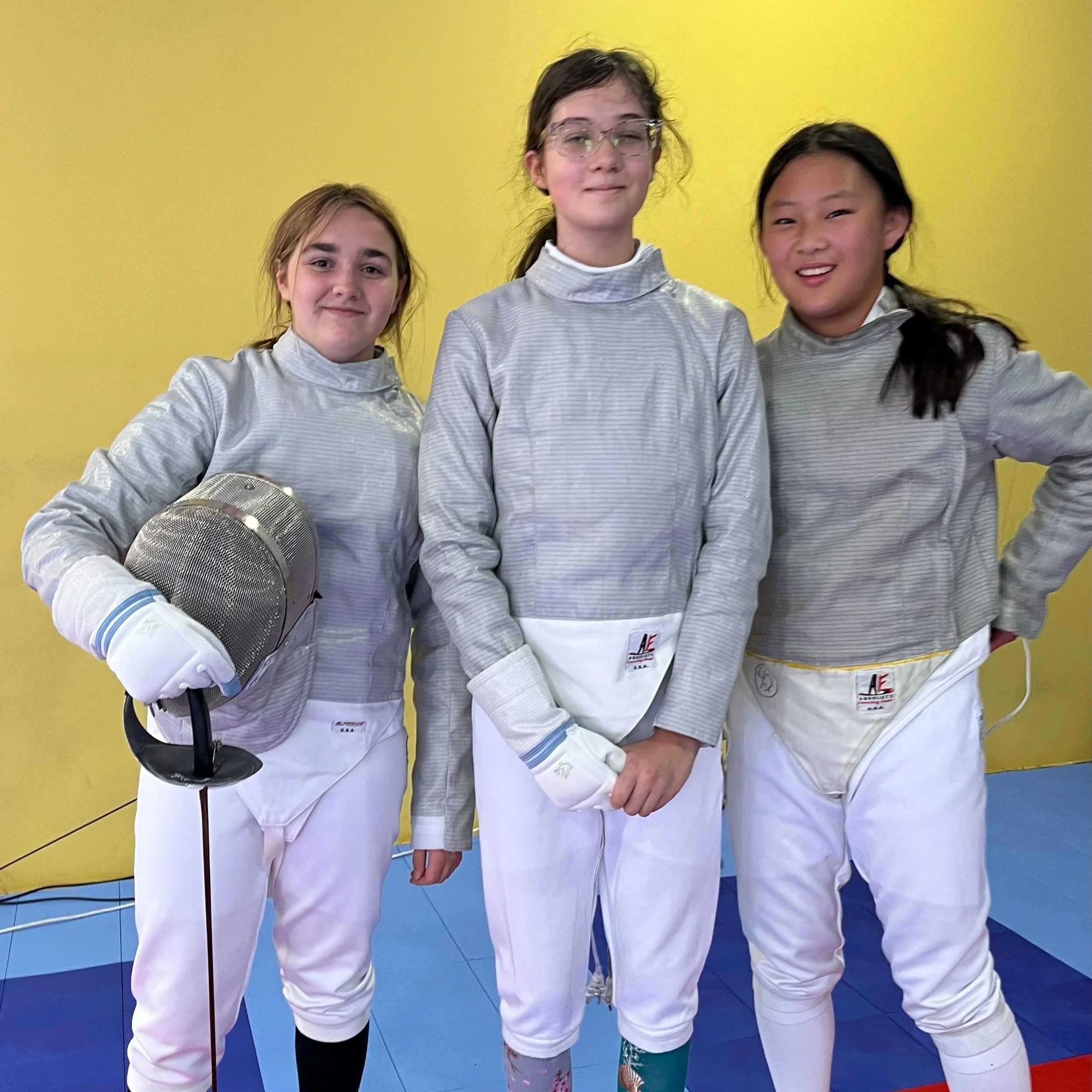 Three saber fencers in fencing gear pose after a tournament at Fence St. Louis fencing club. The wall behind them is yellow and the floor is blue.