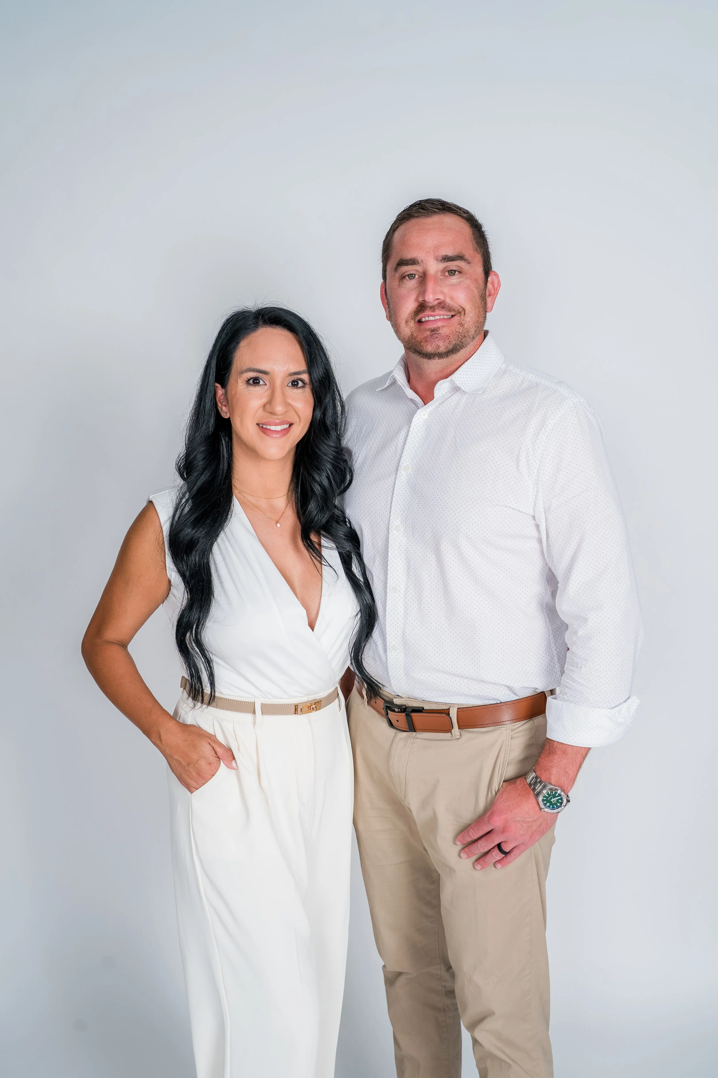 A man and woman standing back to back against a plain white background, looking at the camera. The woman has long dark hair, is wearing gray scrubs, and has her arms crossed. The man has short dark hair, a beard, and is wearing a black polo shirt and gray pants with his hands in his pockets.