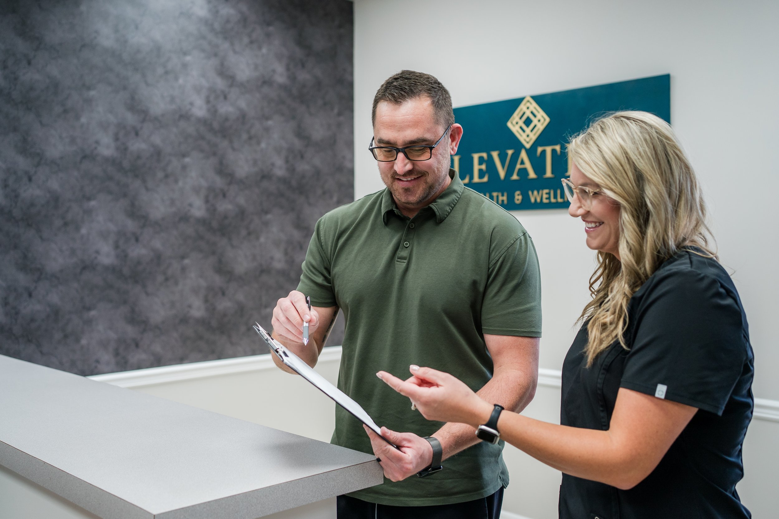 A man and woman standing at a reception desk, smiling and looking at a clipboard together, in a wellness center with a sign that reads 'Elevation Health & Wellness' on the wall behind them.