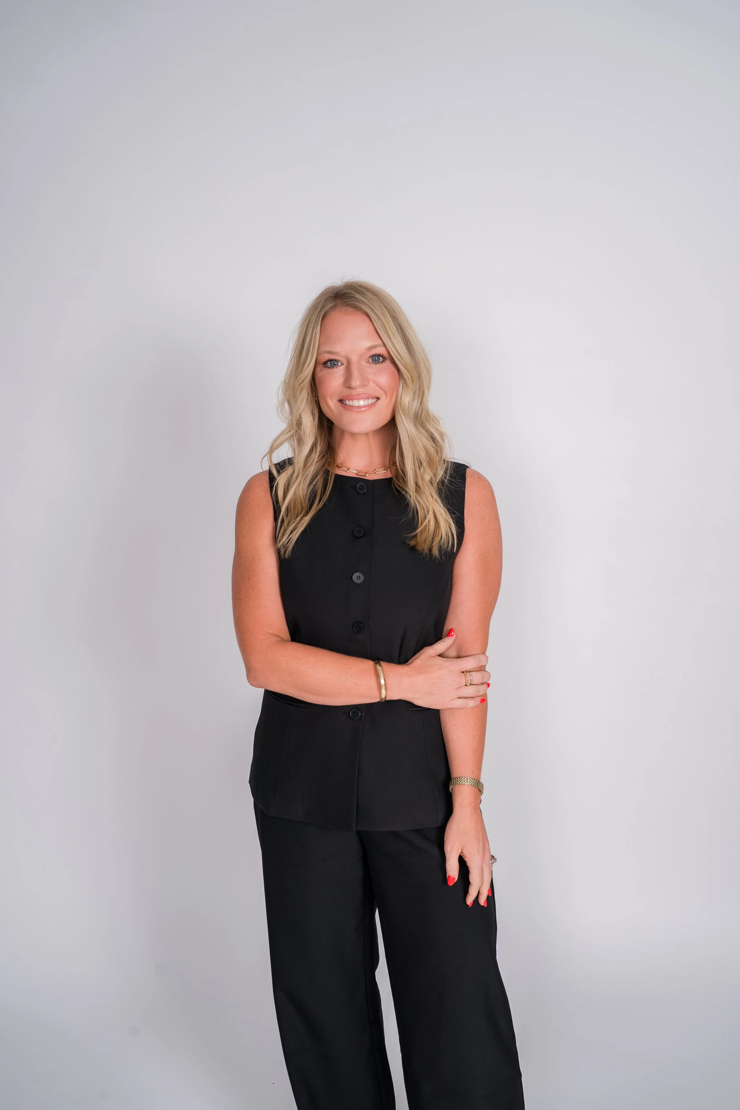 A smiling blonde woman with wavy hair wearing a black top, standing against a gray background.