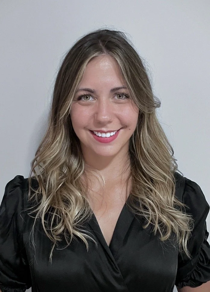 A woman with blonde, wavy hair smiling at the camera, wearing a black top, standing against a plain light-colored wall.