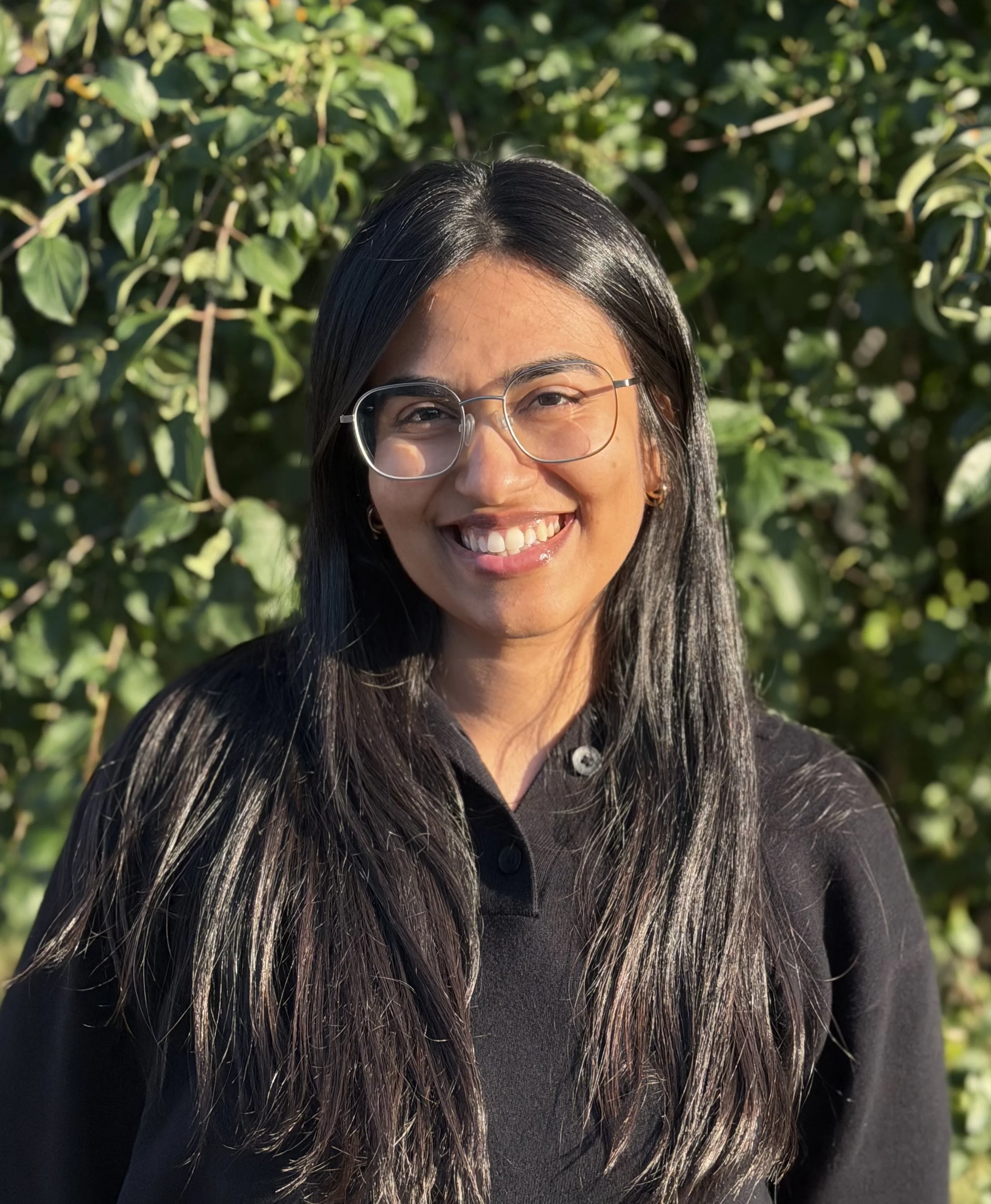 Portrait of a young woman with shoulder-length brown hair, wearing large round glasses and a black sweater, smiling against a plain light-colored wall.