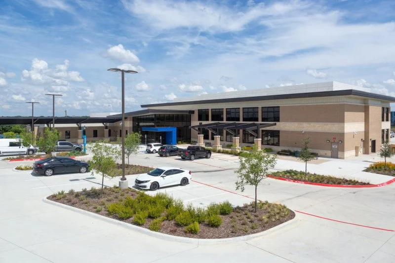 A modern outpatient clinic or medical facility with a parking lot, several parked cars, and landscaping, under a partly cloudy sky.