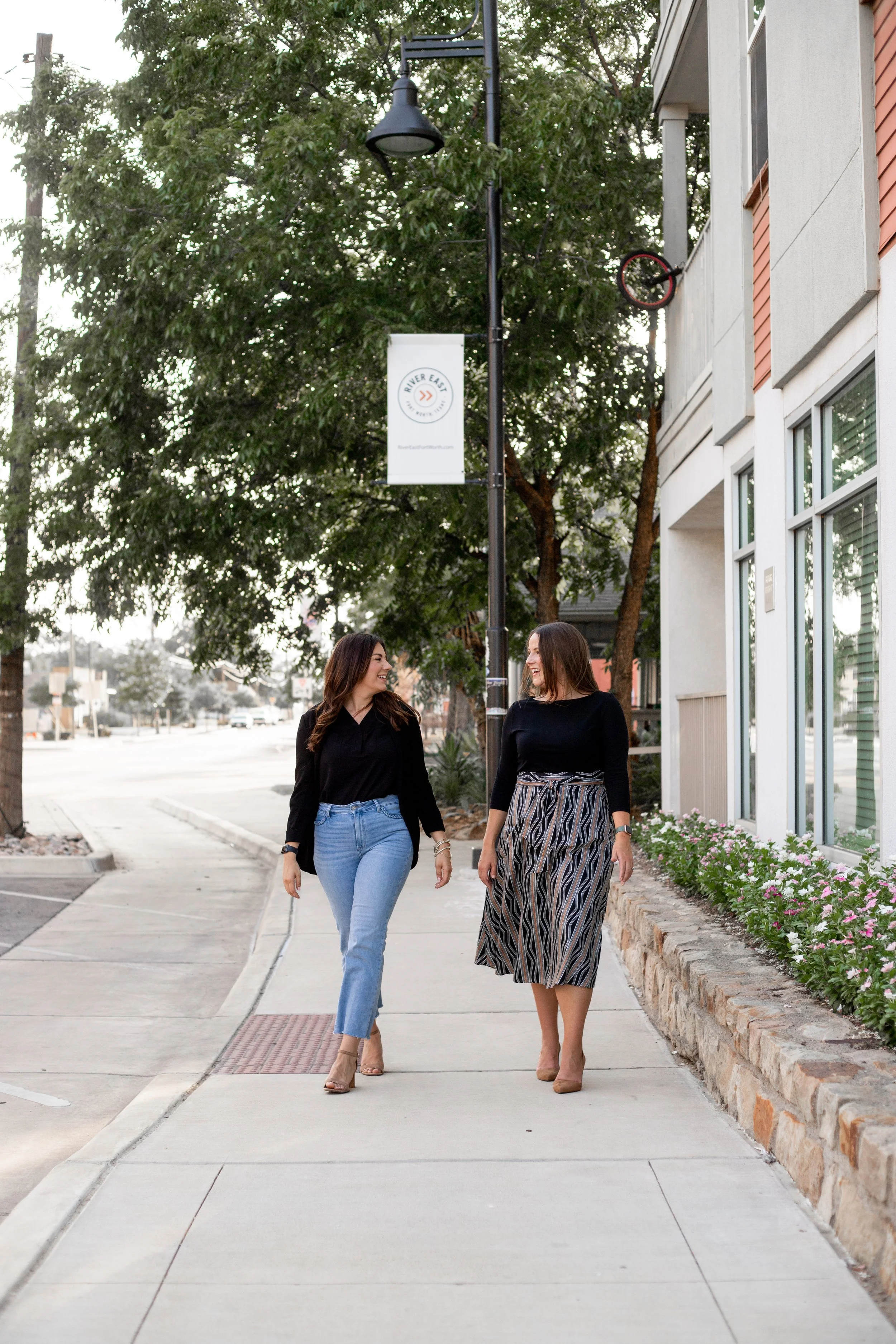 Two women walking and talking on a sidewalk in a suburban area with shops, trees, and flowers.