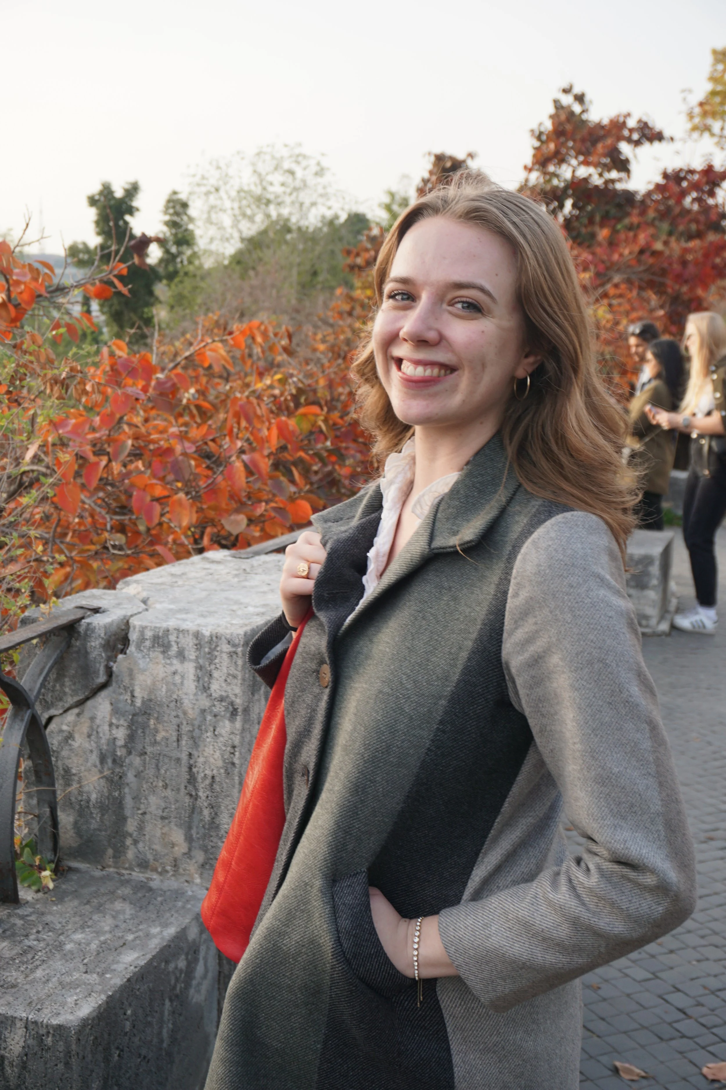 A young woman with light skin and brown hair smiling at the camera, holding her coat, standing outdoors with autumnal foliage and a few people in the background.