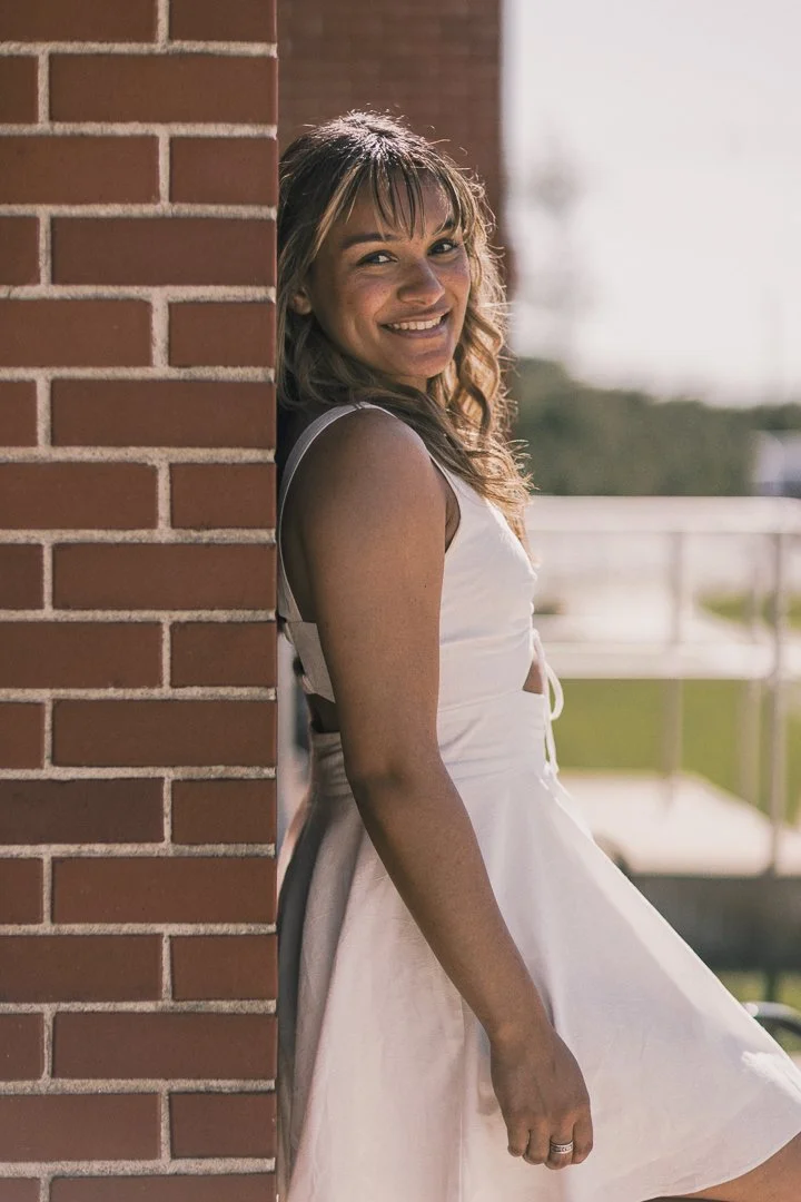 A smiling woman in a white dress leaning against a brick wall outdoors on a sunny day.