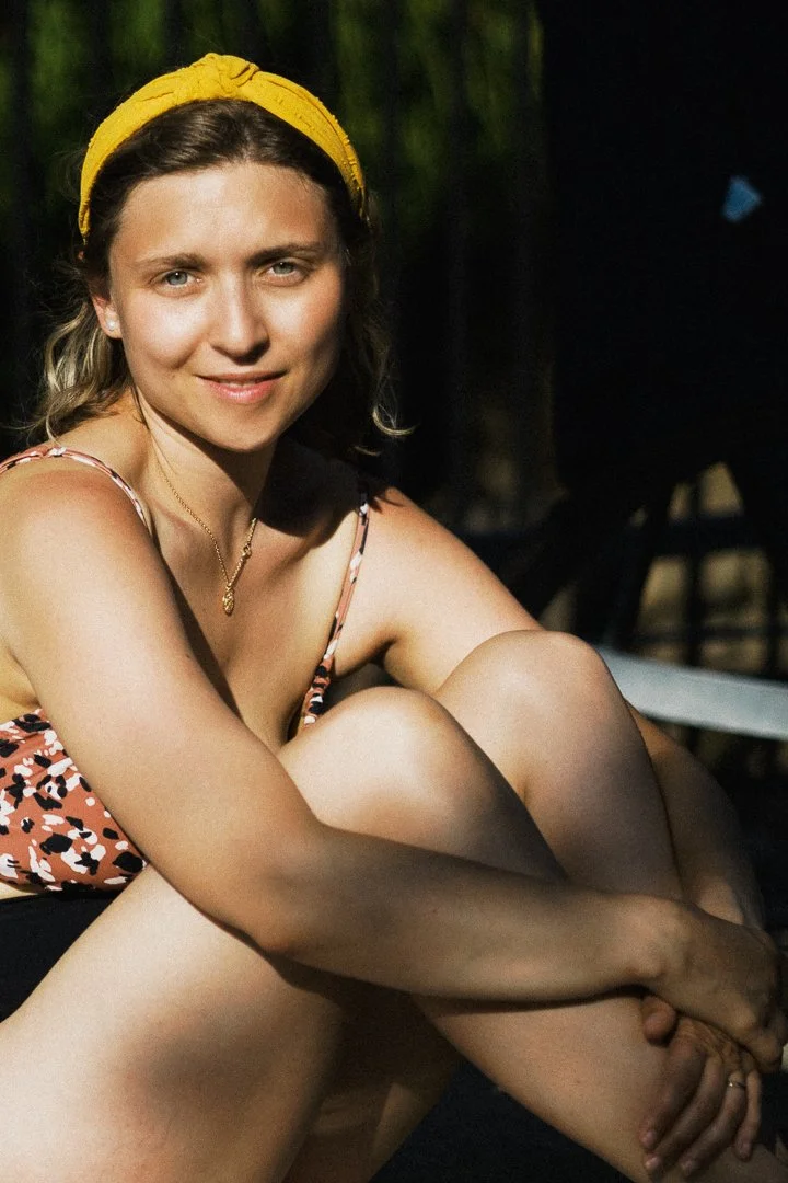 A woman with a yellow headband, wearing a patterned swimsuit, sitting outdoors and smiling at the camera.