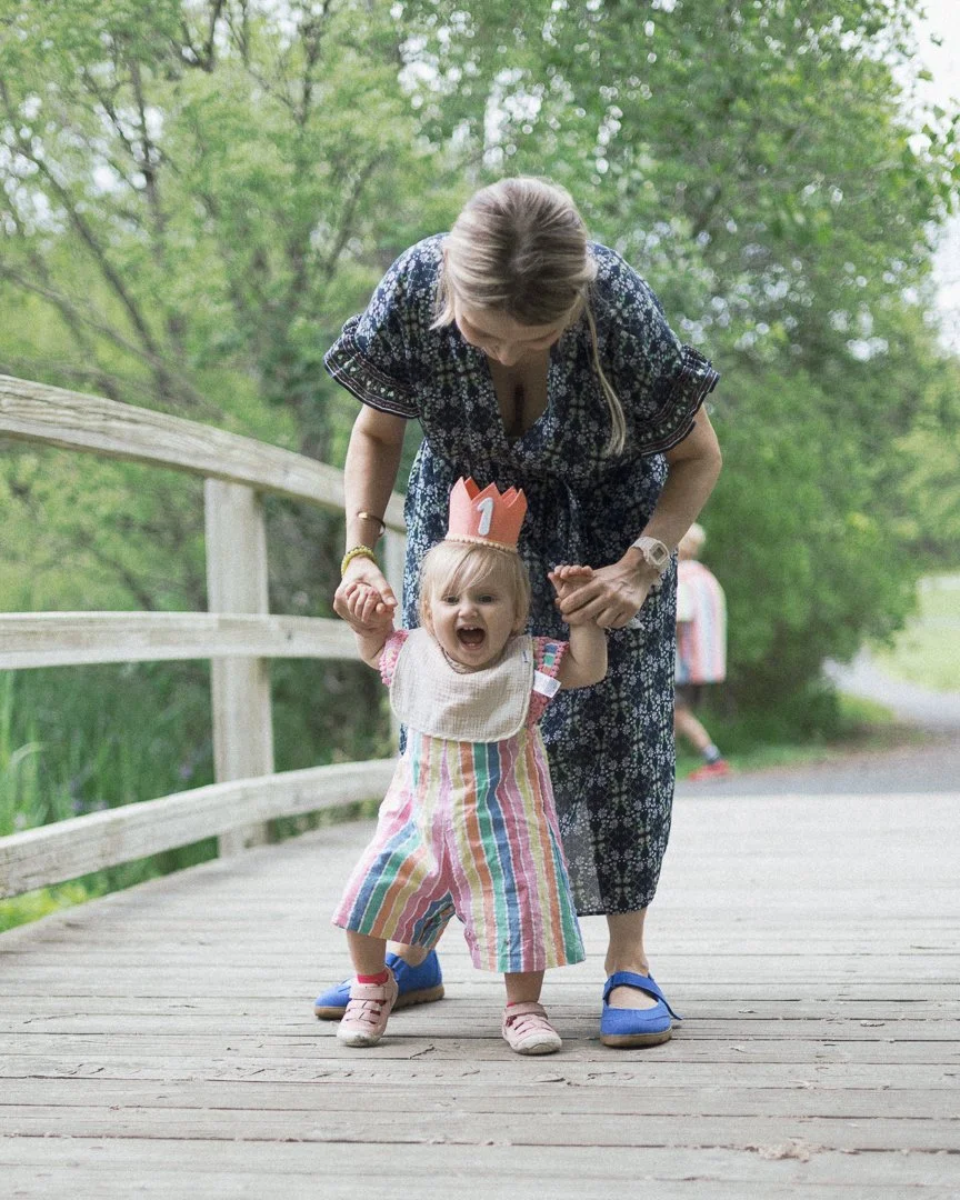 A young girl with a striped dress and a birthday crown is walking on a wooden bridge, supported by an adult woman.
