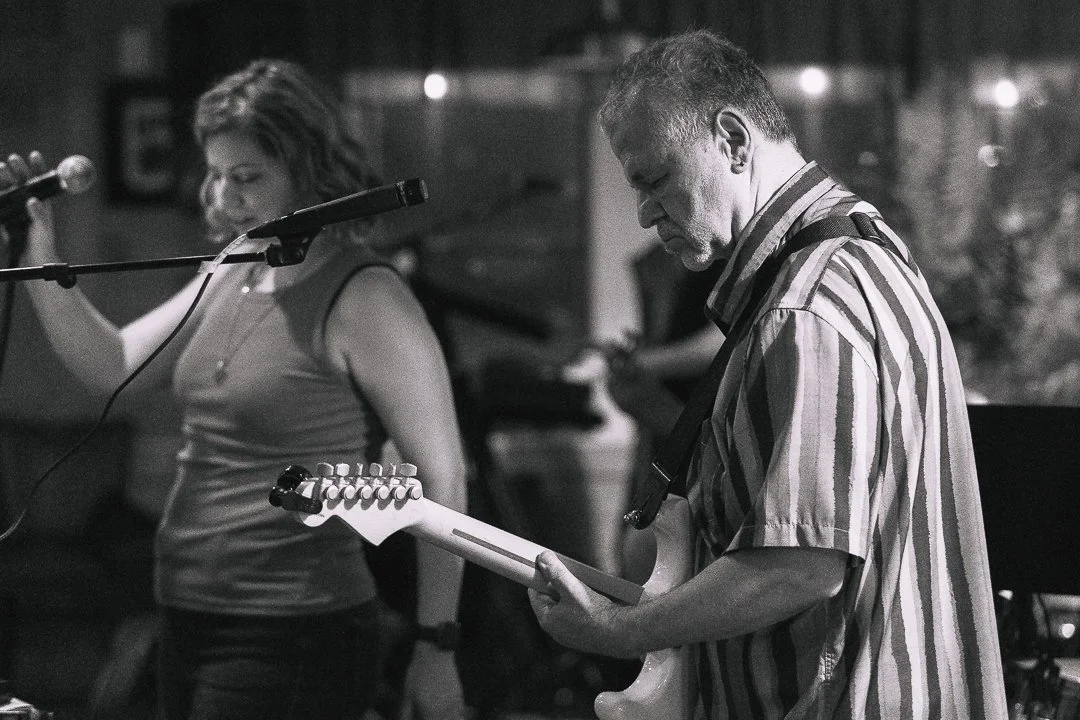 A man playing an electric guitar and a woman standing behind a microphone in a dimly lit setting.