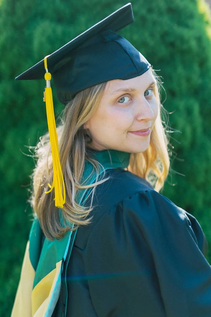 A young woman in a graduation cap and gown smiling over her shoulder outdoors.