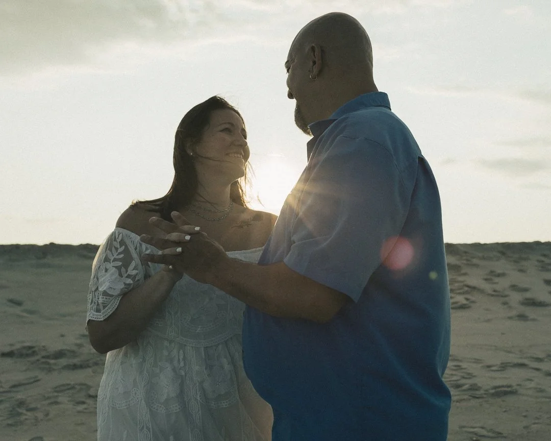 A couple dancing on the beach during sunset, with the woman smiling at the man.