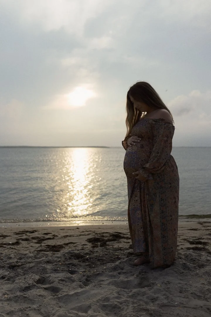 A pregnant woman in a patterned dress standing on a sandy beach during sunset, with her head bowed and arms crossed over her belly.