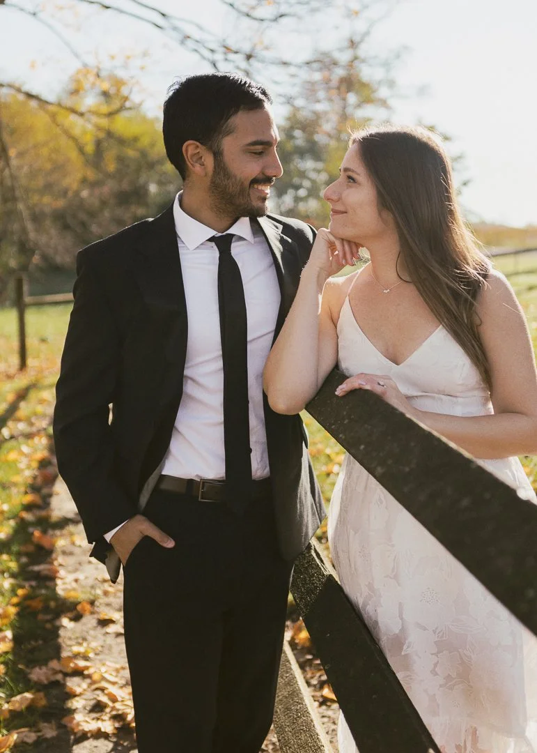 A couple standing outdoors in fall, smiling at each other. The man is dressed in a black suit with a white shirt and black tie, and the woman wears a white dress. They are leaning on a wooden fence near a path with autumn leaves, with trees and sunli