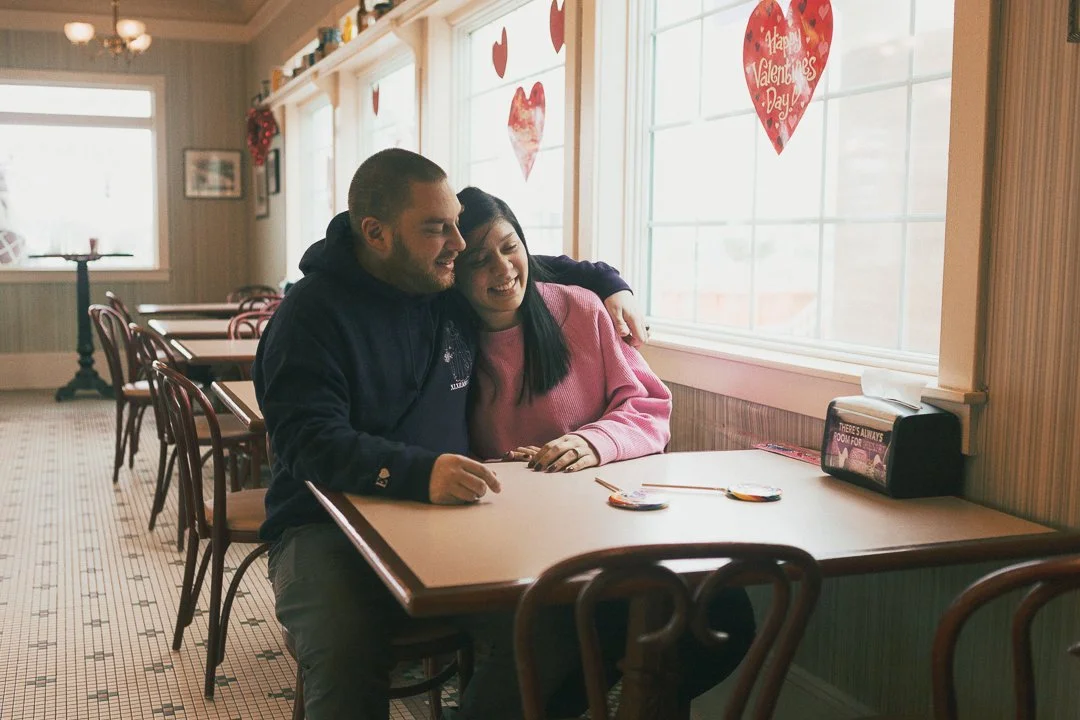 A couple sitting at a restaurant table near a window, hugging and smiling, Valentine's Day decorations on the windows, including a large heart with the message 'Happy Valentine's Day'.
