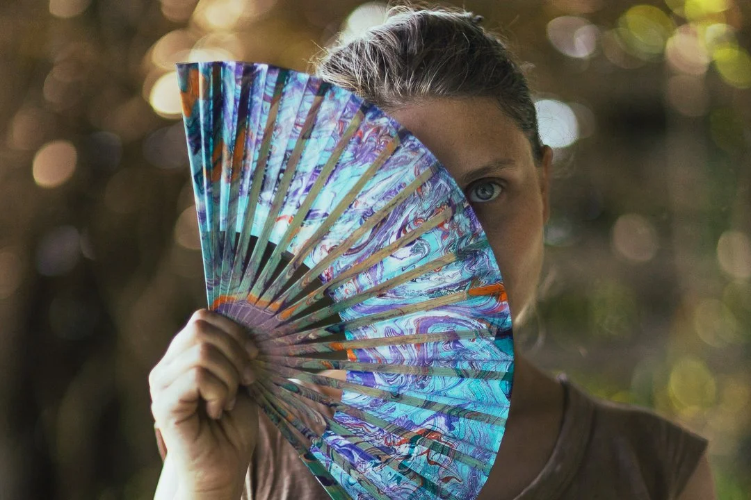 A young woman with blue eyes holds a colorful folding fan partially covering her face, with a blurred outdoor background and bokeh light effects.